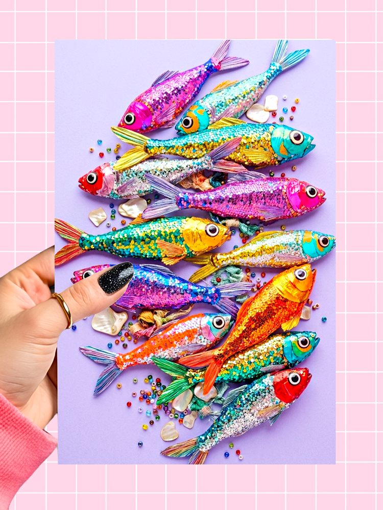 A hand with black glitter nail polish holds a square card featuring colorful, glittery fish-shaped candies scattered on a lavender background, set against a pink grid backdrop.