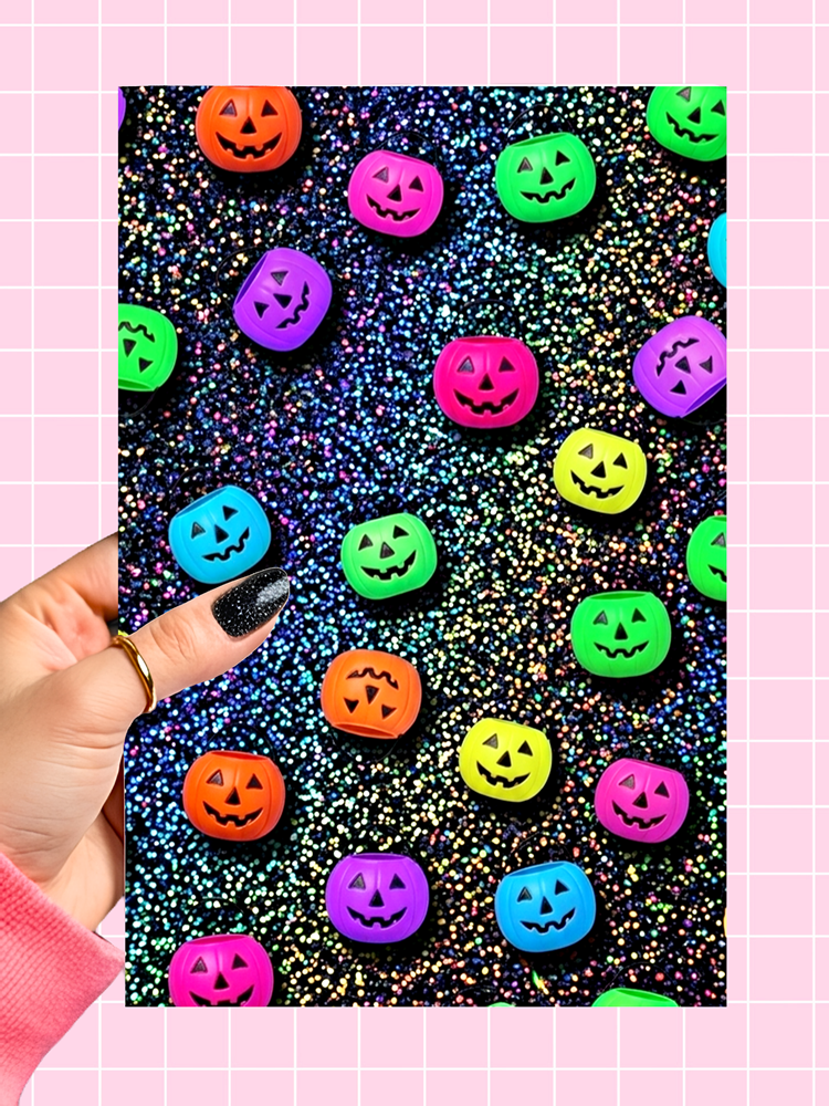 A hand with black glitter nail polish holds a square card showing colorful jack-o'-lantern candies on a black glitter background, set against a pink grid backdrop.