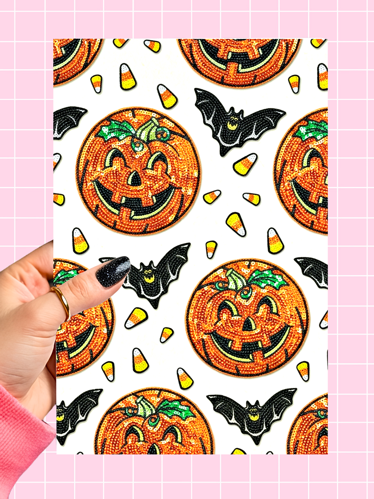 A hand with black glitter nail polish holds a rectangular sheet featuring a repeating Halloween pattern of orange sequined jack-o'-lanterns, black bats, and candy corn, set against a pink grid background.
