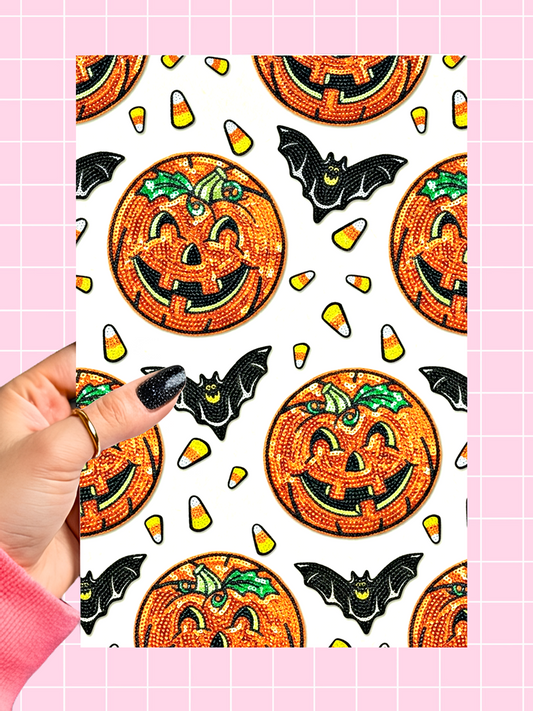 A hand with black glitter nail polish holds a rectangular sheet featuring a repeating Halloween pattern of orange sequined jack-o'-lanterns, black bats, and candy corn, set against a pink grid background.