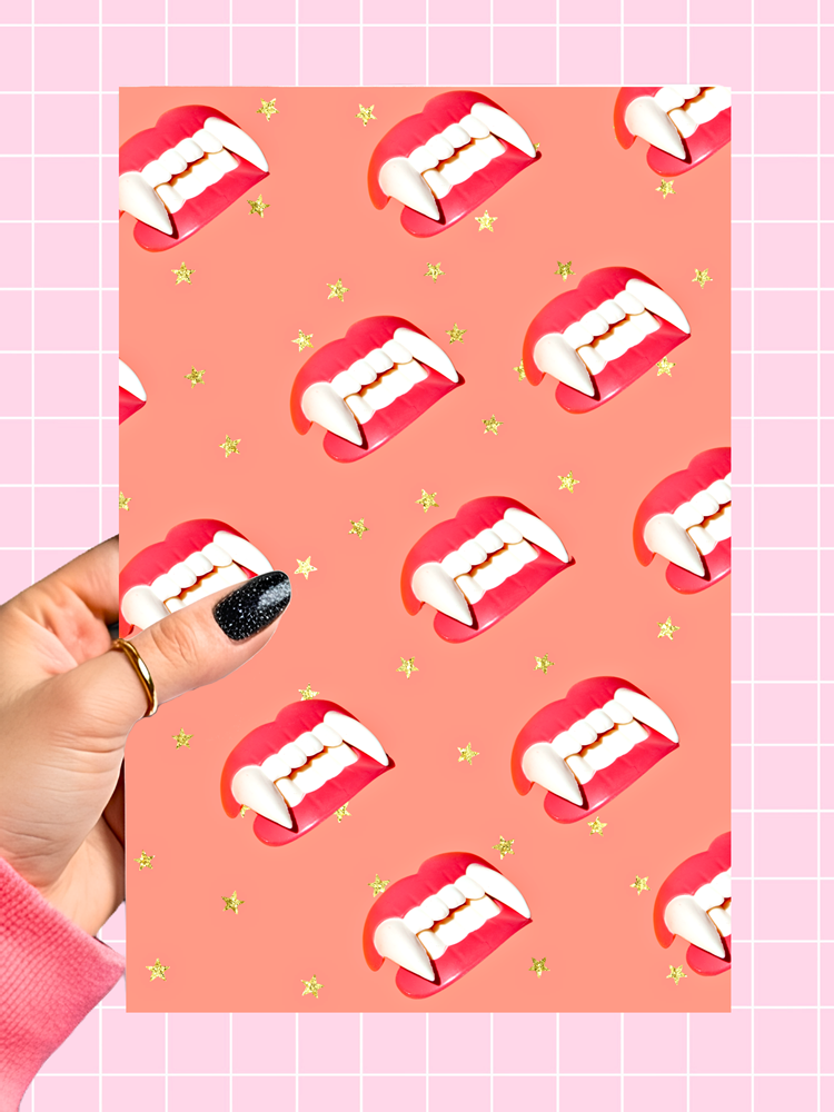 A hand with black glitter nail polish holds a rectangular card featuring a repeating pattern of vampire fangs in red and white, set against a peach background with small gold stars. The card is positioned over a pink grid background.