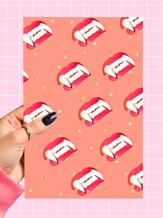 A hand with black glitter nail polish holds a rectangular card featuring a repeating pattern of vampire fangs in red and white, set against a peach background with small gold stars. The card is positioned over a pink grid background.