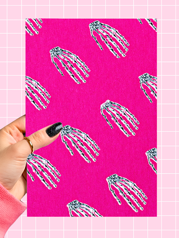 A hand with black glitter nail polish and a gold ring holds a pink canvas print featuring a repeating pattern of white and black skeletal hands. The background is a light pink grid.