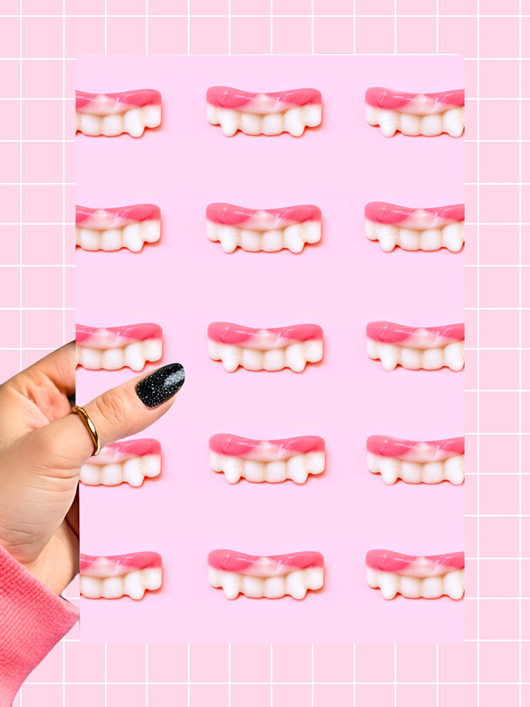 A hand with black glitter nail polish holds a sheet of pink gummy candies shaped like vampire teeth against a pink grid background.