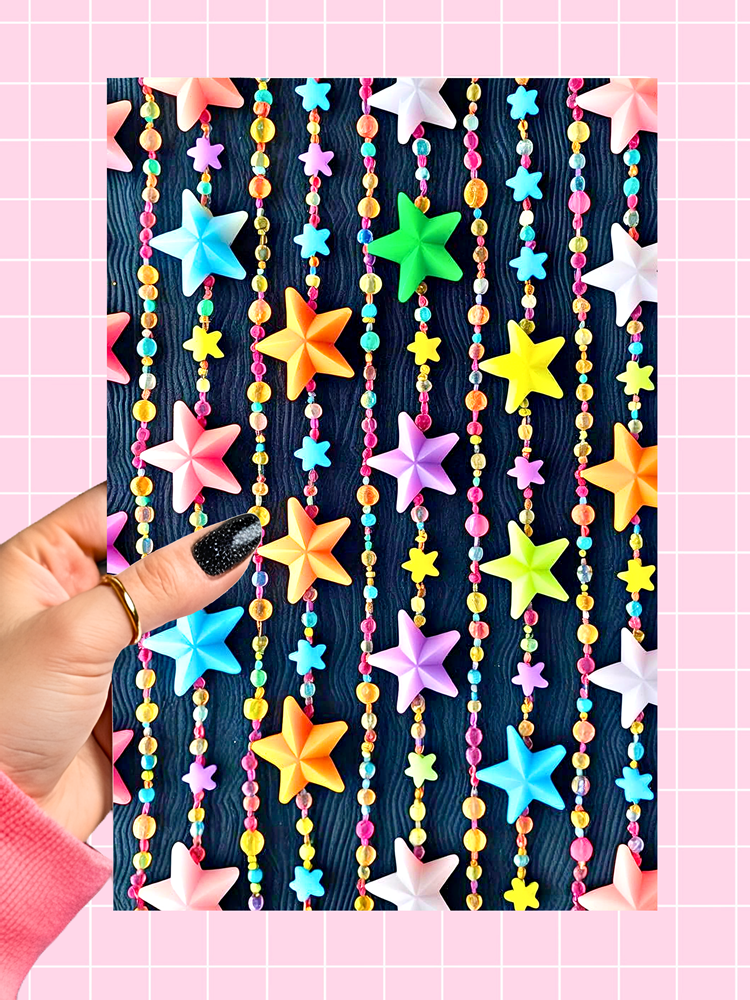 A hand with black glitter nail polish holds a decorative card featuring colorful star-shaped beads and hanging beaded strands on a dark wood-grain background, set against a pink grid backdrop.