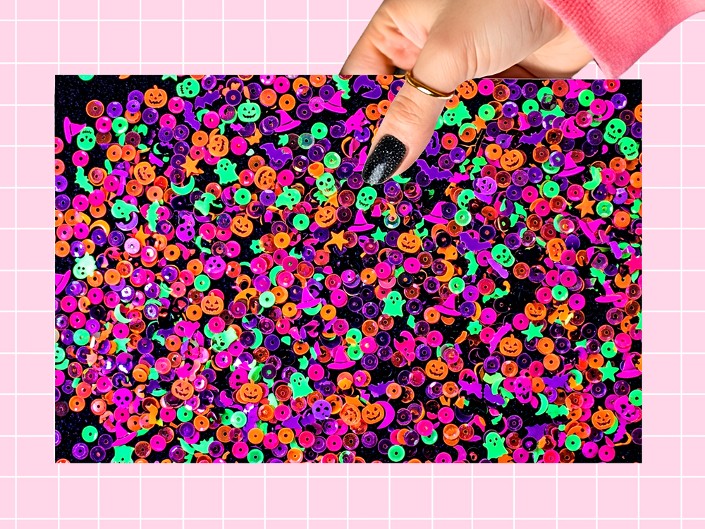 A hand with black glitter nail polish holds a rectangular sheet of colorful Halloween sequins featuring jack-o'-lanterns, ghosts, witches, and moons on a black background, set against a pink grid backdrop.