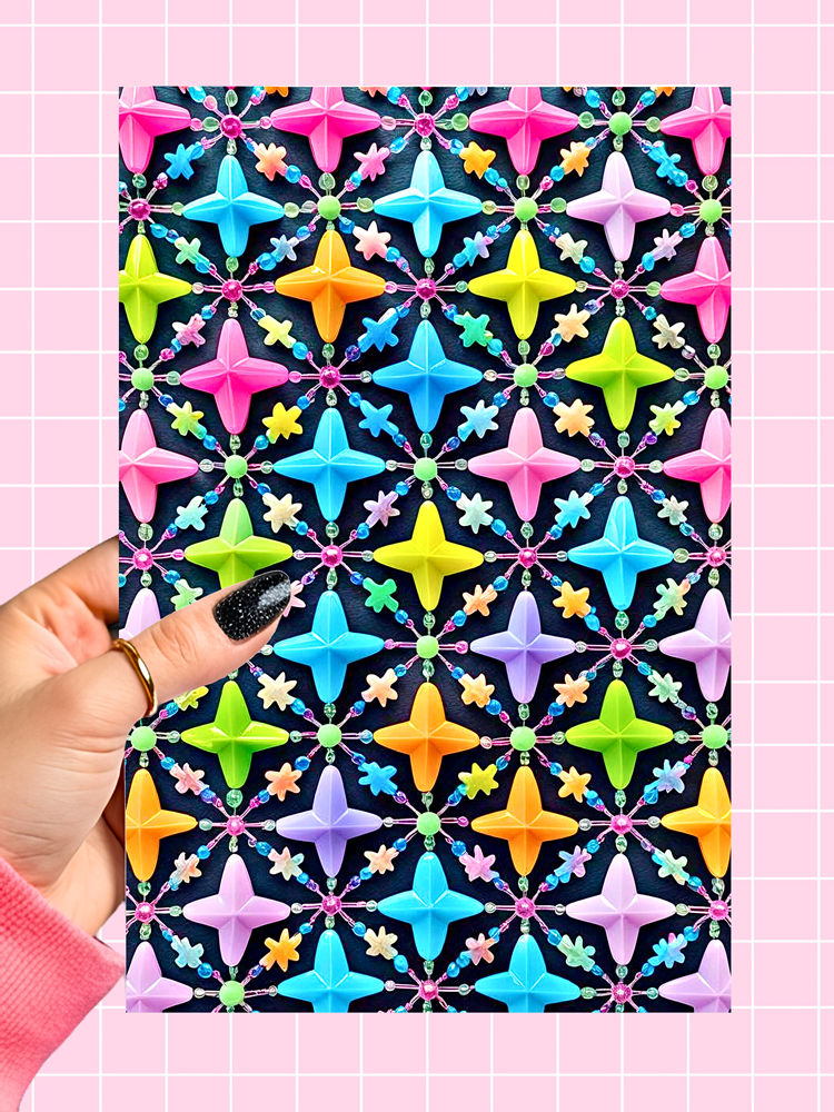 A hand with black glitter nail polish holds a square card or print featuring a colorful geometric pattern of star-like shapes on a dark background. The print is set against a light pink grid backdrop.