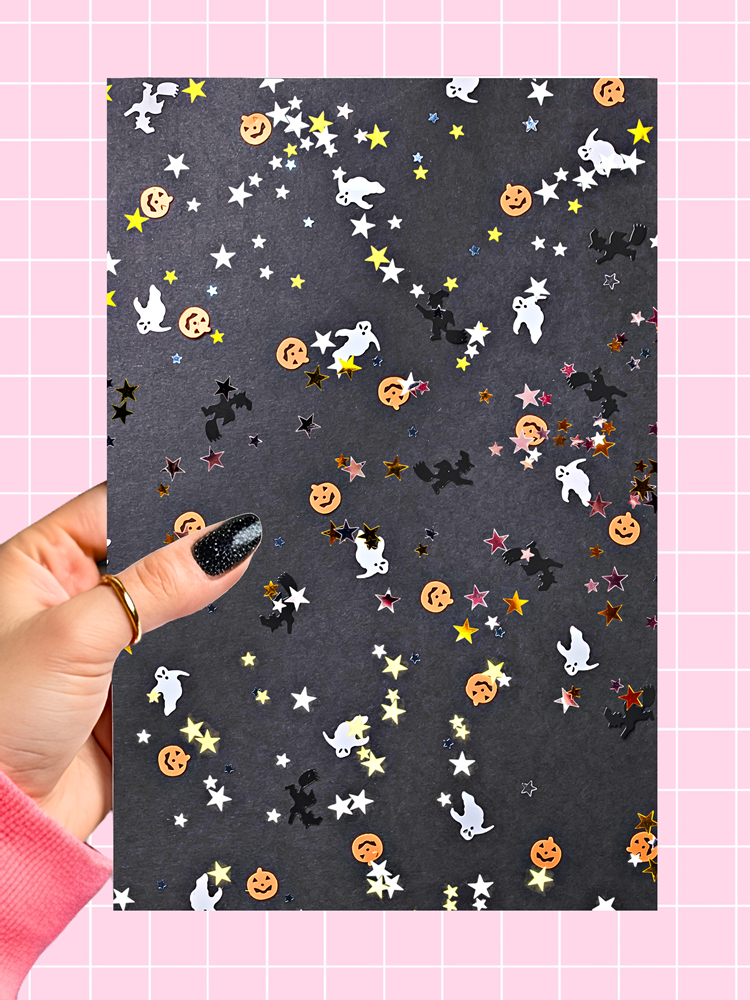 A hand with glittery black nail polish and a gold ring holds a sheet of black craft paper decorated with colorful Halloween confetti, including white ghosts, orange pumpkins, black bats, and silver stars, against a pink grid background.