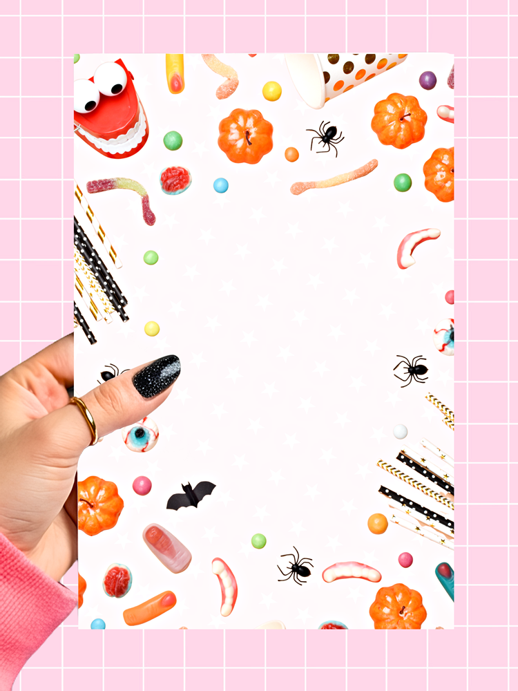 A hand with black glitter nail polish holds a white notepad decorated with Halloween-themed illustrations like candy corn, gummy worms, bats, spiders, and ghosts, set against a pink grid background.