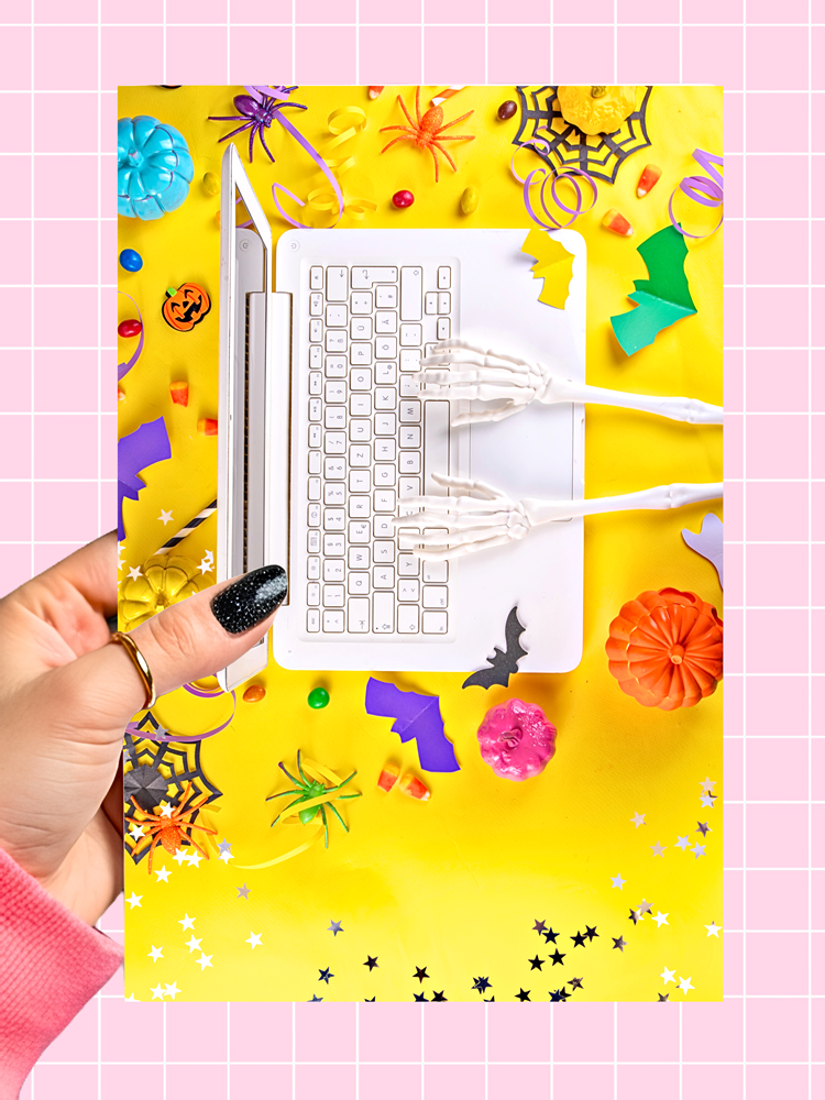 A hand with black glitter nail polish holds a printed card featuring a white laptop surrounded by skeleton hands, orange pumpkins, spiderwebs, and Halloween decorations on a bright yellow background. The card is set against a pink grid backdrop.