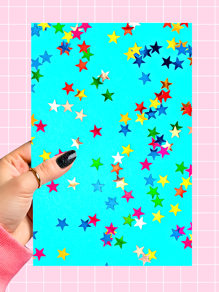 A hand with black glitter nail polish holds a rectangular card featuring a bright turquoise background covered in scattered colorful star cutouts in red, yellow, blue, green, and white, against a pink grid backdrop.