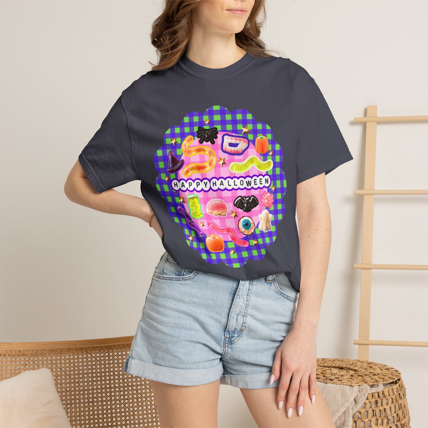 A woman wearing a dark charcoal gray t-shirt with a colorful 'Happy Halloween' graphic print featuring candy and sushi illustrations, paired with light-wash denim shorts in a cozy indoor setting.