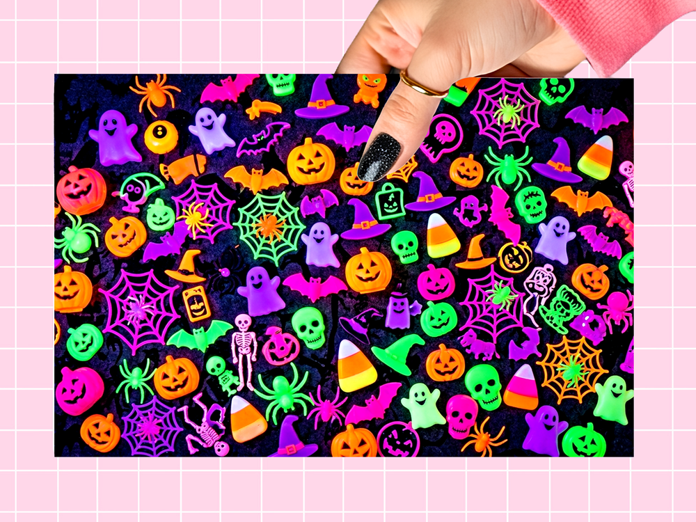 A hand with black glitter nail polish points to a sheet of colorful Halloween-themed glitter stickers on a pink grid background. The stickers feature glowing neon designs including pumpkins, ghosts, spiders, bats, witches hats, and candy corn on a black glitter base.