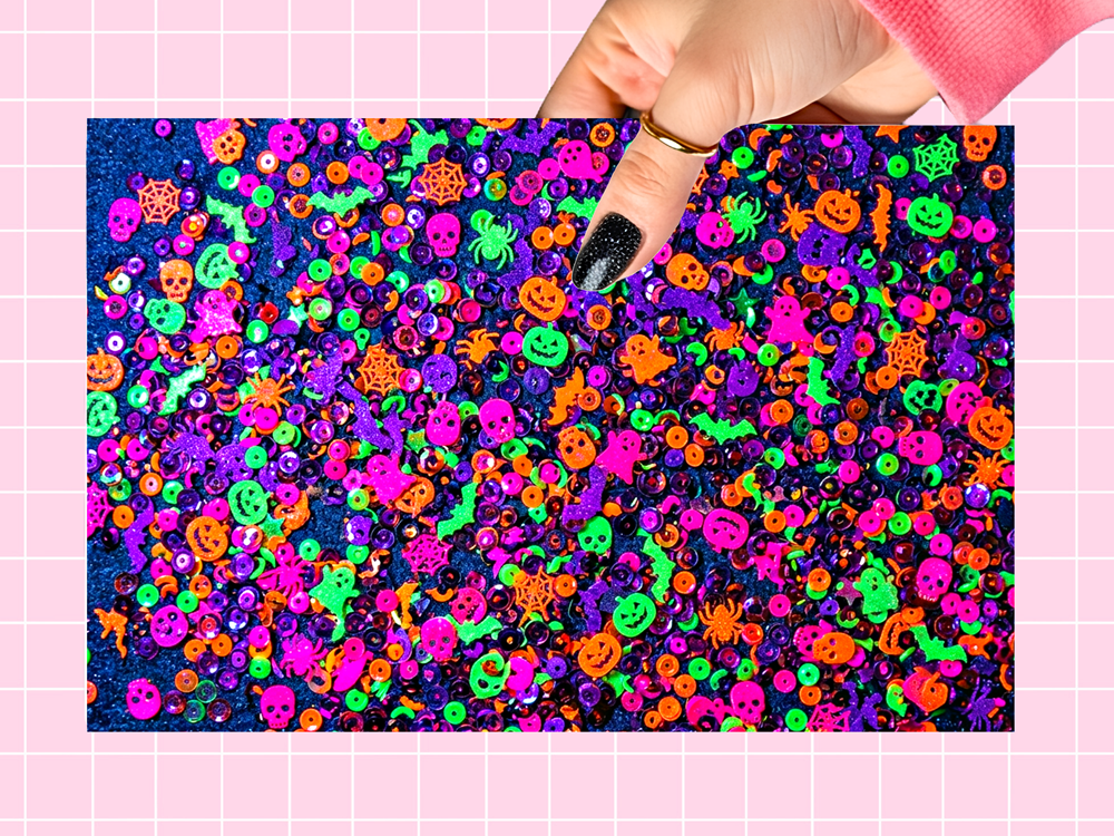 A hand with black glitter nail polish holds a rectangular sheet covered in dense, colorful Halloween-themed sequins and glitter, including pumpkins, spiders, ghosts, and stars in neon pink, green, orange, purple, and blue against a pink grid background.