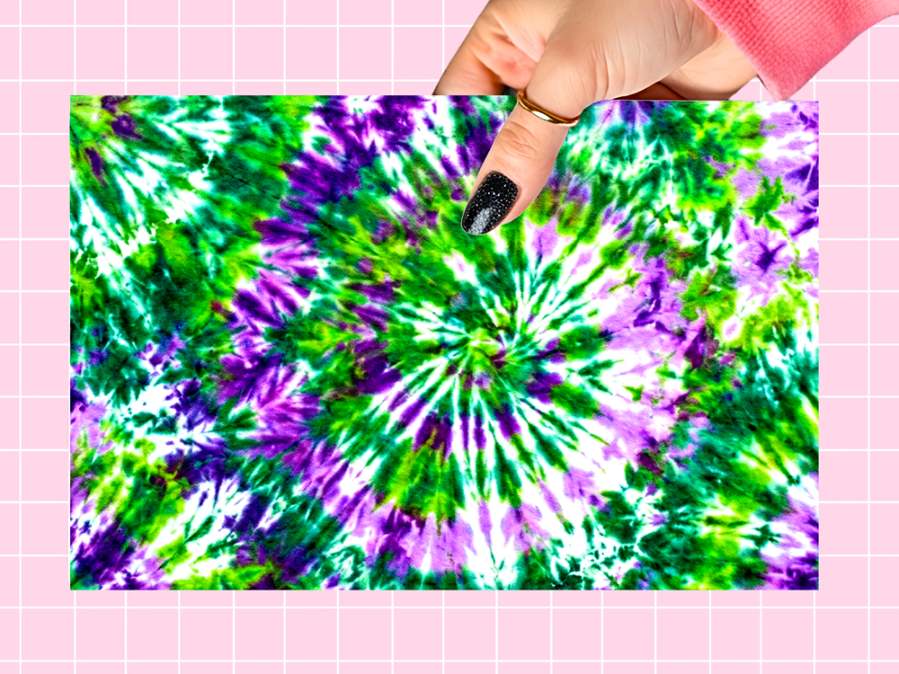 A rectangular fabric or paper sample with a bright tie-dye spiral pattern in green, purple, and white, held by a hand against a pink grid background.