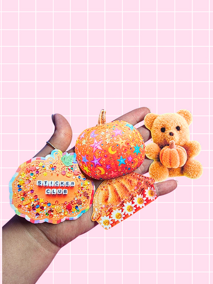 A hand holding three decorative autumn-themed stickers against a pink grid background: a pumpkin-shaped sticker with stars and moons, a round sticker reading 'STICKER CLUB' with colorful confetti, and a small plush teddy bear holding a pumpkin.