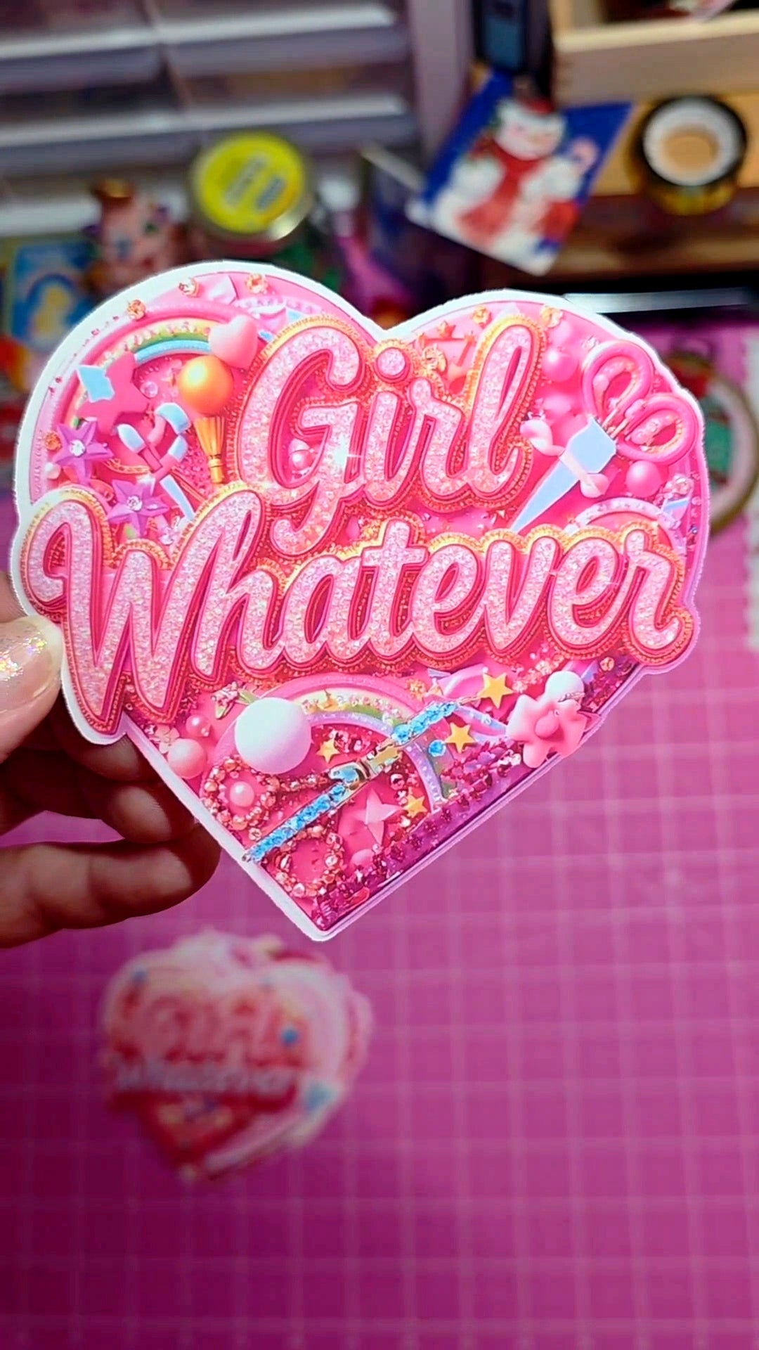 Heart-shaped sticker with 'Girl Whatever' text held against a pink background