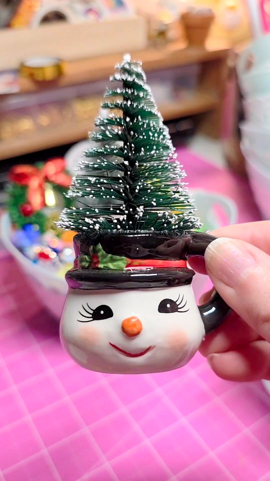 Snowman-shaped mug with a Christmas tree on top, held by a hand against a blurred background.