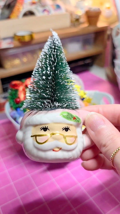 Small Santa Claus figurine with a Christmas tree on top, held by a hand against a colorful background.
