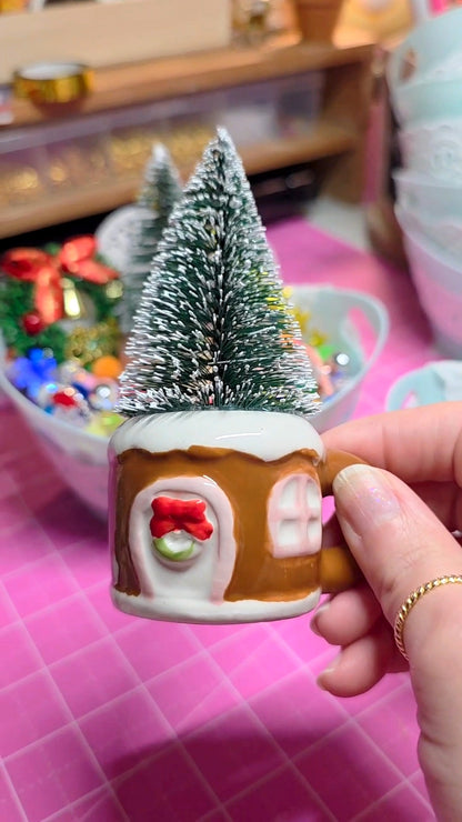 Small decorative Christmas tree in a gingerbread house-shaped container held by a hand on a pink surface.