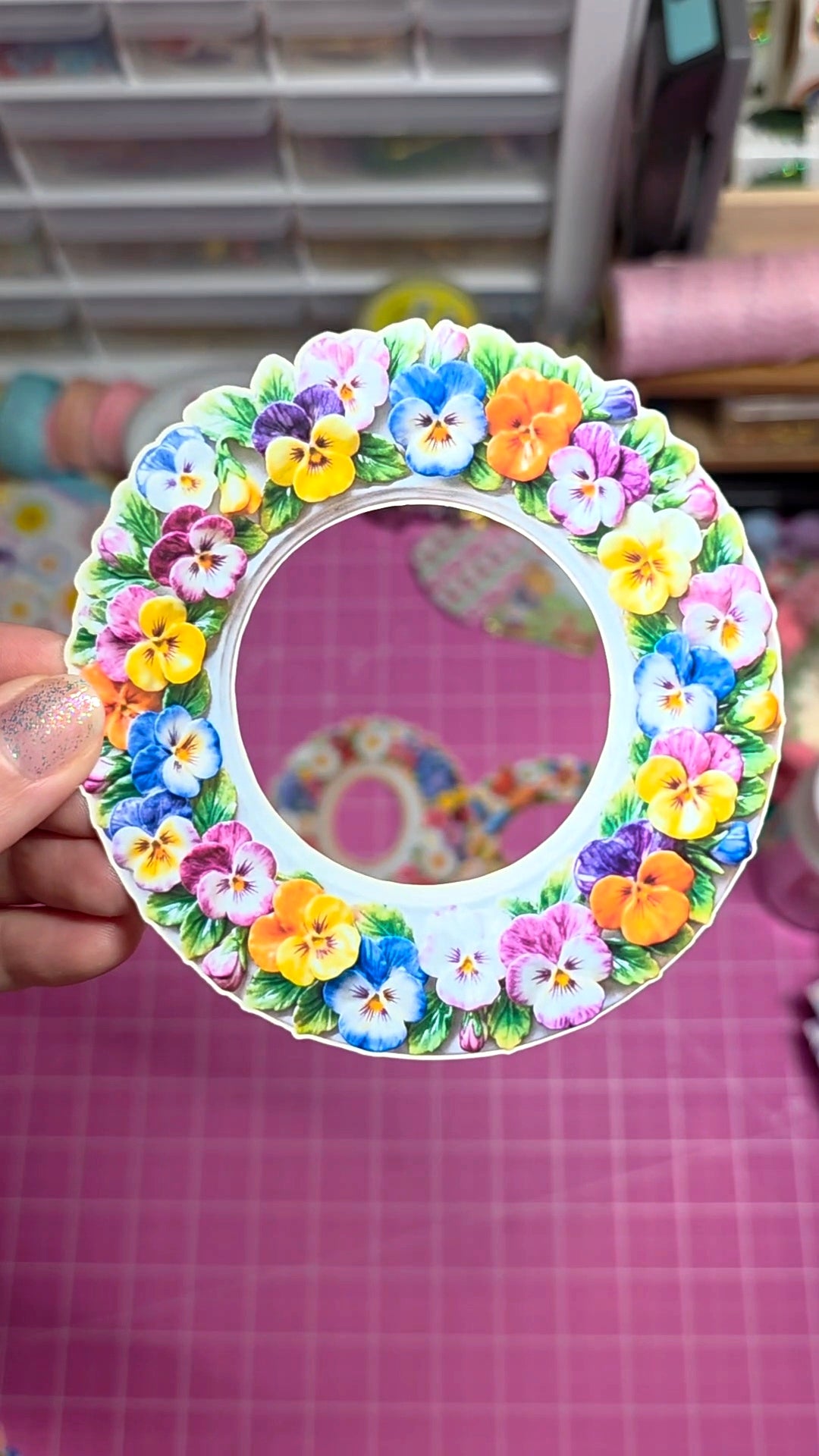 Circular mirror with colorful floral design held by a hand on a pink grid background.