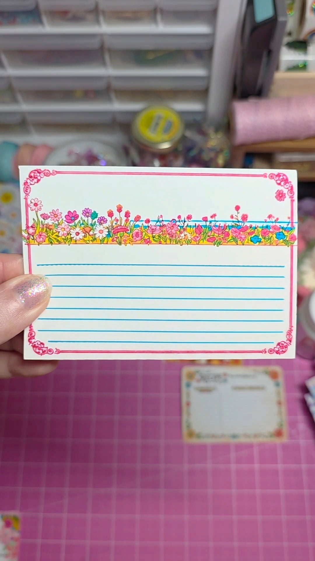 Colorful stationery item with decorative border held by a hand on a pink grid background.