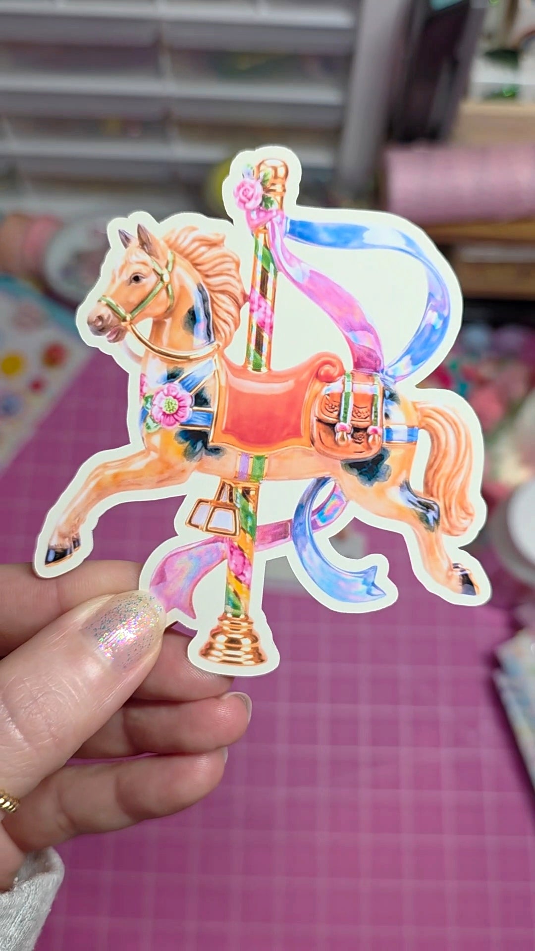 Colorful carousel horse sticker held by a hand on a pink grid background