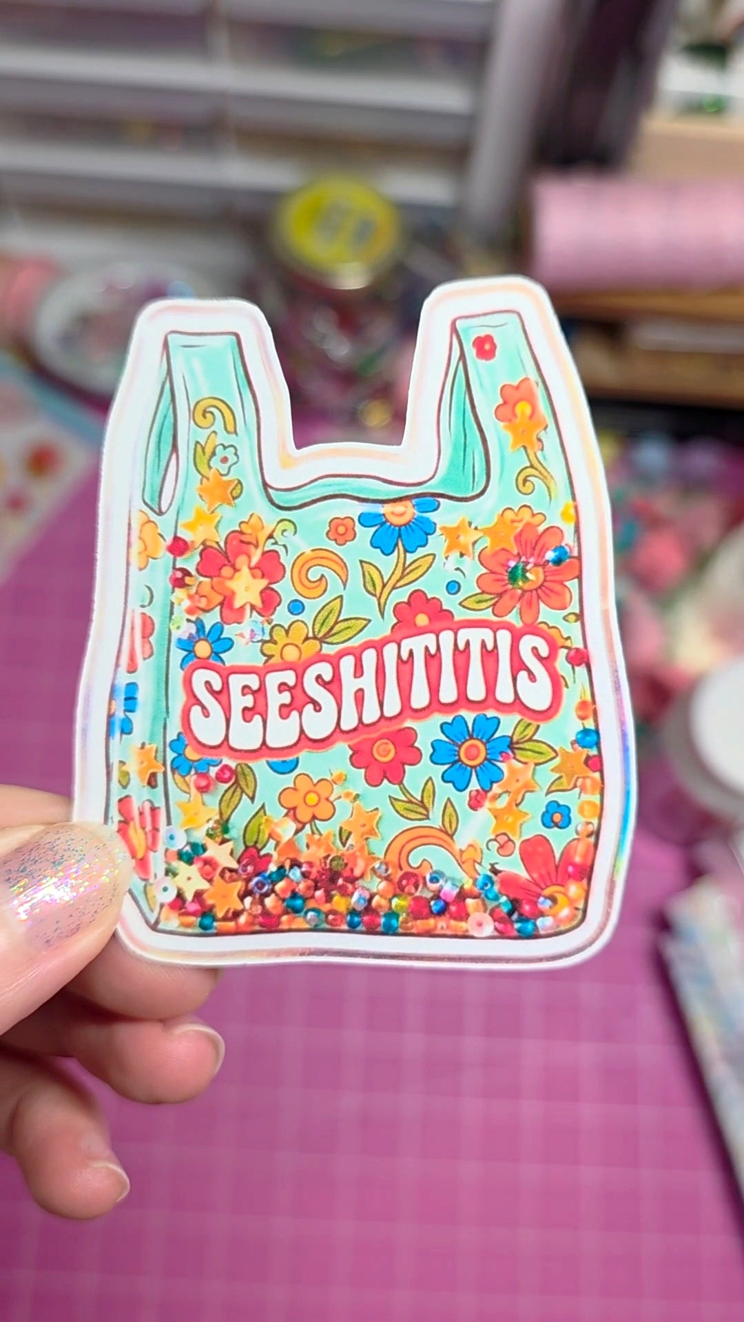 Colorful sticker with floral design and 'SEESHITITIS' text held by a hand on a pink background.