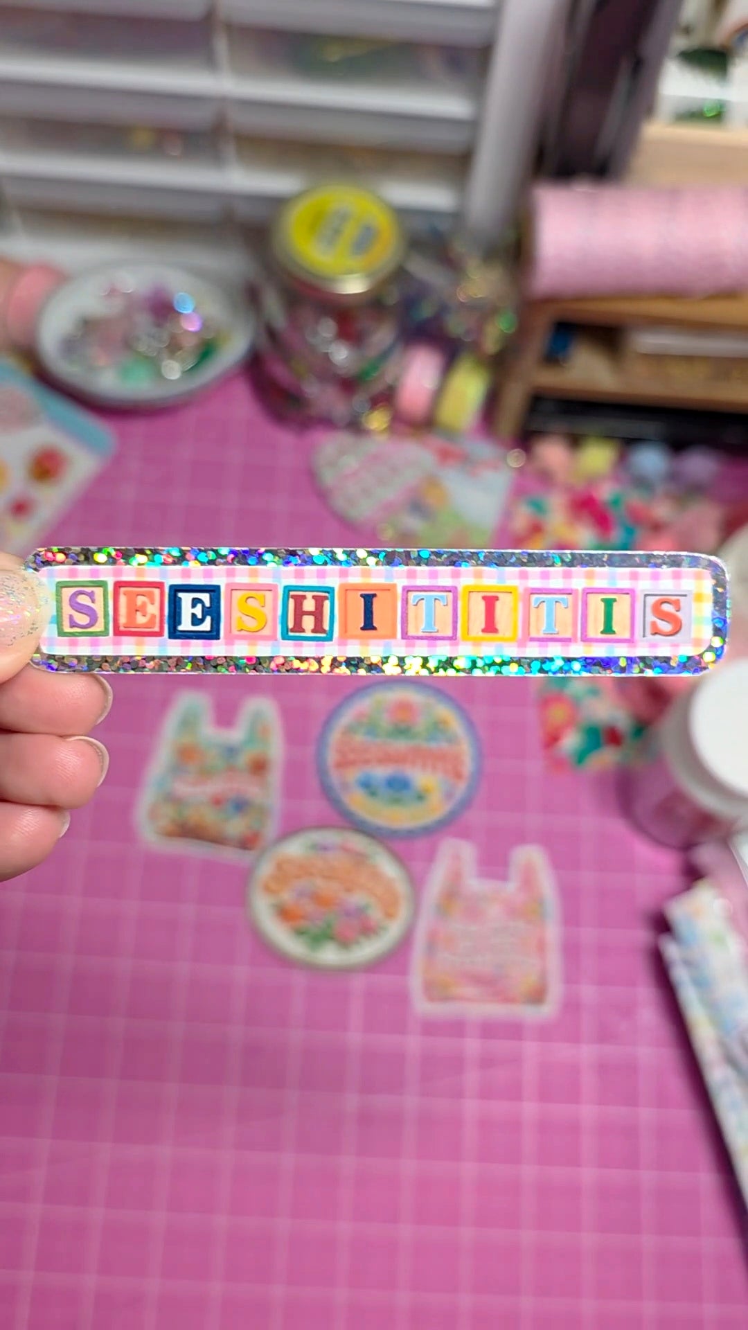 Colorful rectangular sticker with 'SEESHITITIS' text held over a pink surface with stationery items.