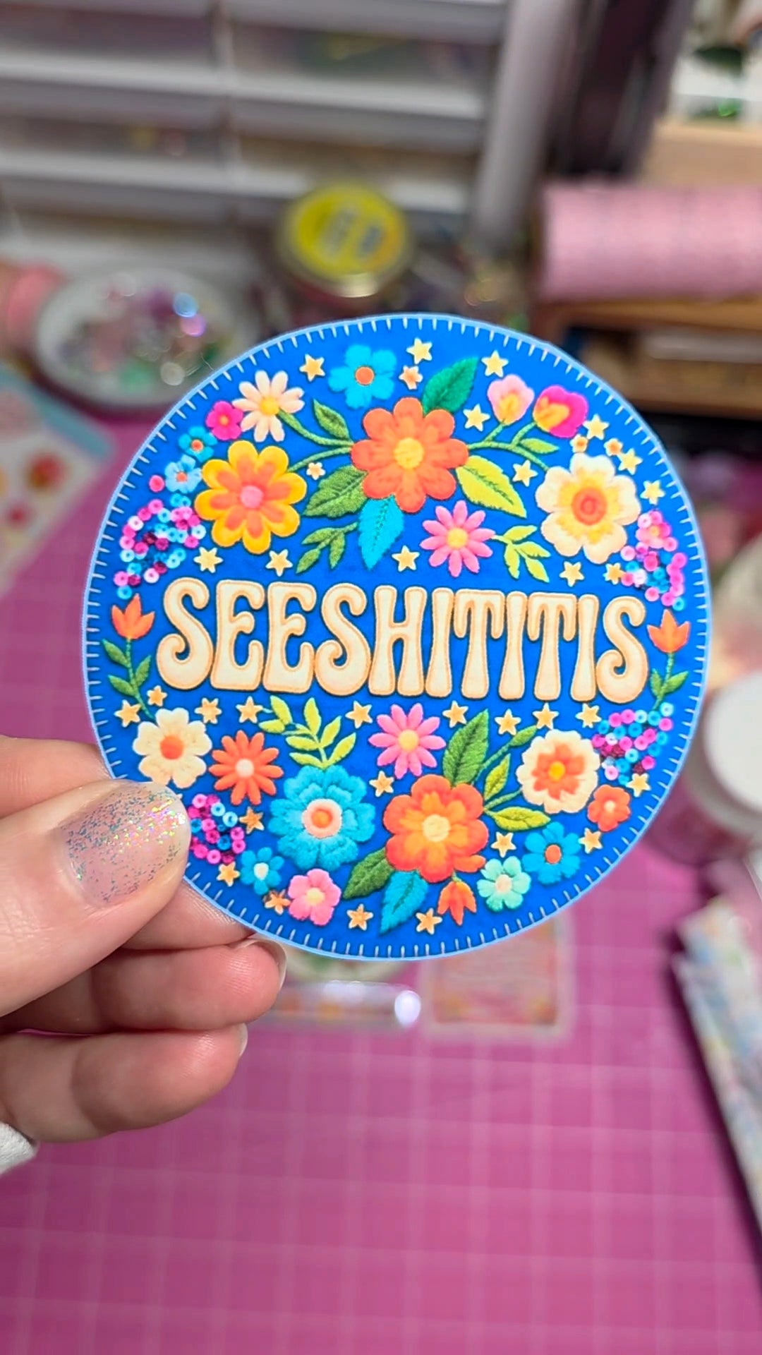 Colorful sticker with floral design and 'SEESHITTIES' text held by a hand on a pink surface.