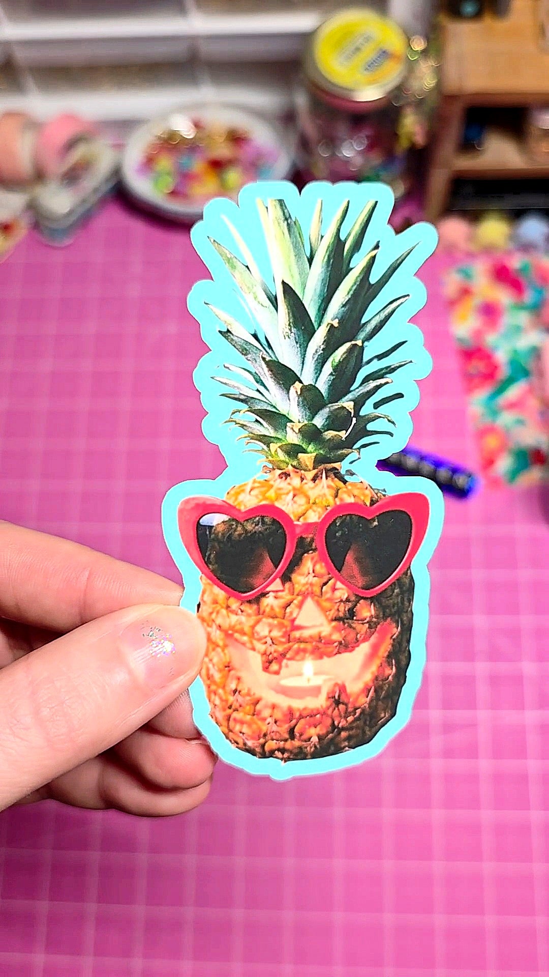 Sticker of a pineapple wearing sunglasses held by a hand on a pink checkered background.
