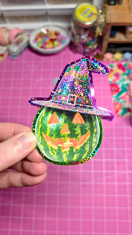 Decorative watermelon with a witch hat and carved face held by a hand on a pink grid background.