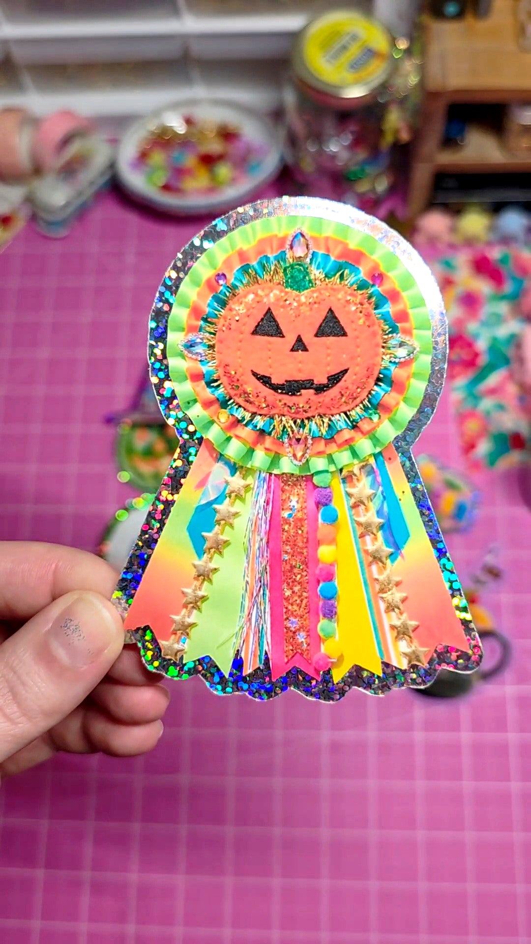 Colorful Halloween-themed ribbon with a jack-o'-lantern face held by a hand on a pink grid background.
