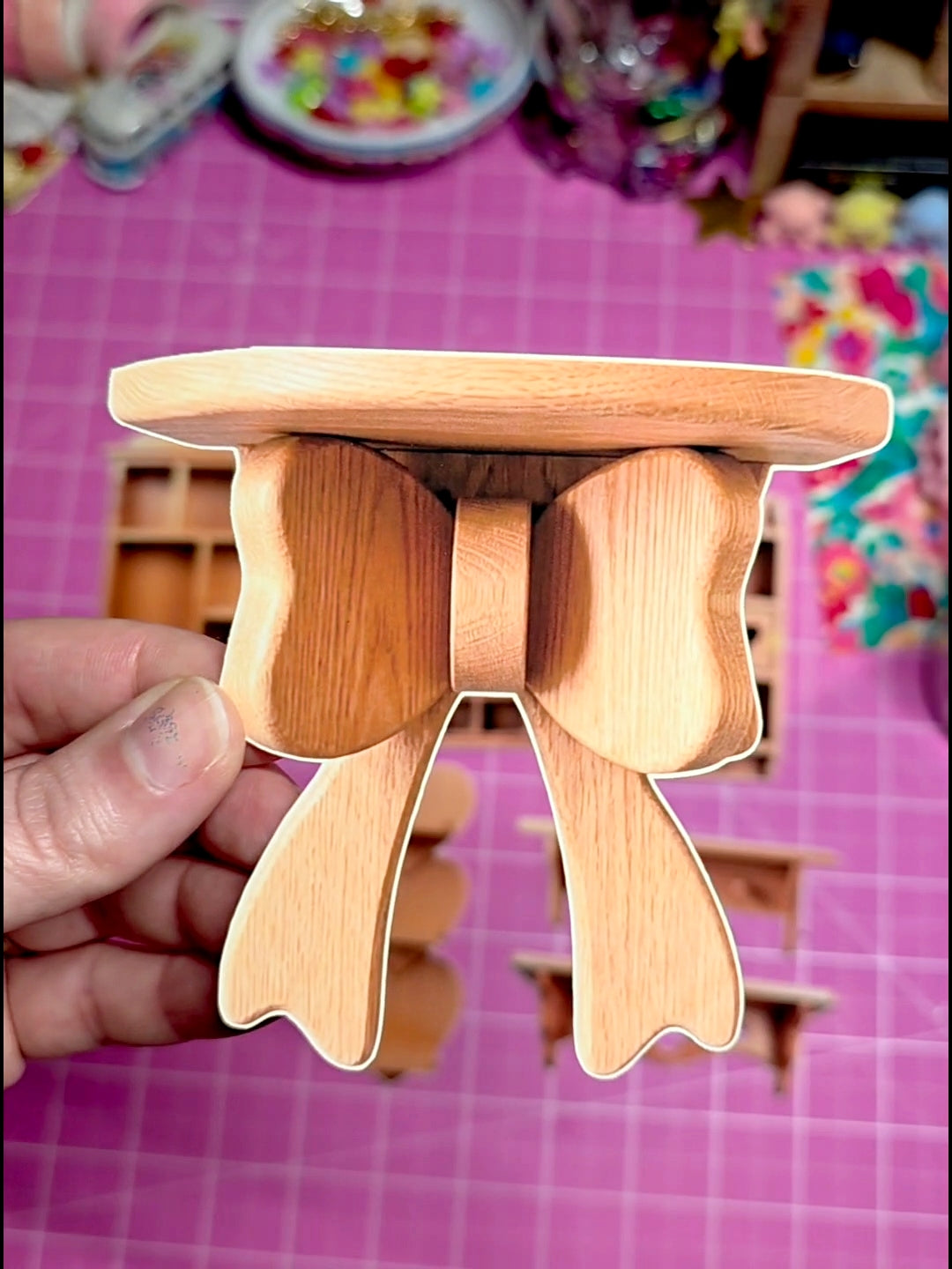 Wooden stool with a small round platform held by a hand on a pink grid background.