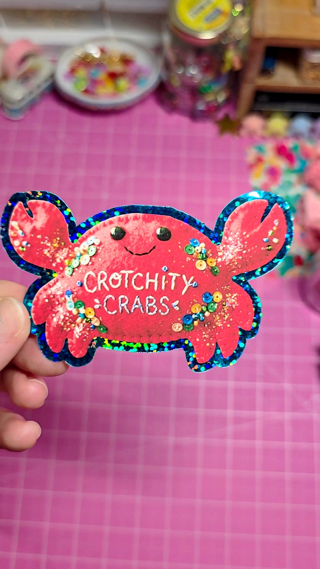 Pink crab-shaped sticker with 'Crotchity Crabs' text held by a hand on a pink grid background.