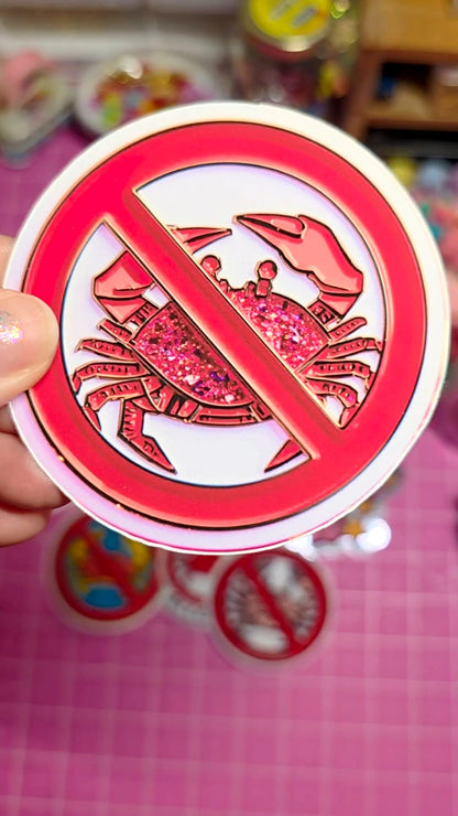 Circular sticker with a red prohibition symbol over a crab illustration, held against a blurred background.